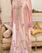 A light pink traditional long dress with purple and green floral embroidery, shown on a mannequin in an elegant cream-colored room with a sofa in the background.