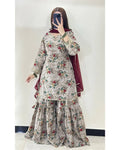 A woman takes a mirror selfie wearing a traditional three-piece salwar kameez set. The set features a light beige kurta and flared pants with a red, green, and blue floral pattern, paired with a deep maroon dupatta.