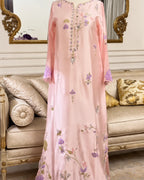 A long pink embroidered dress or caftan with floral and geometric motifs in purple, green, and gold, displayed on a mannequin in front of an ornate beige sofa and mirror.