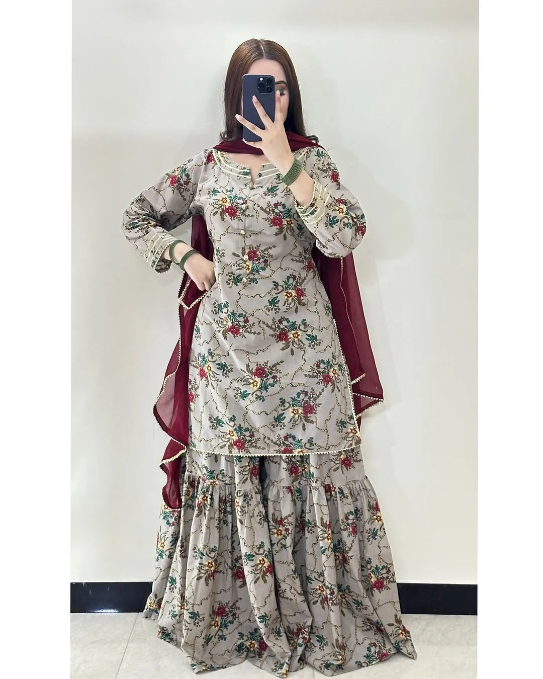 A woman modeling a traditional Indian salwar kameez suit with a beige floral print, a deep maroon dupatta, and a matching maroon inner skirt, photographed in a mirror selfie style against a plain indoor wall.