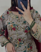 A close-up mirror selfie of a person wearing a beige embroidered kurta with a red floral pattern, green and gold accents, and a dark red dupatta. The person is holding a dark blue smartphone to take the photo.
