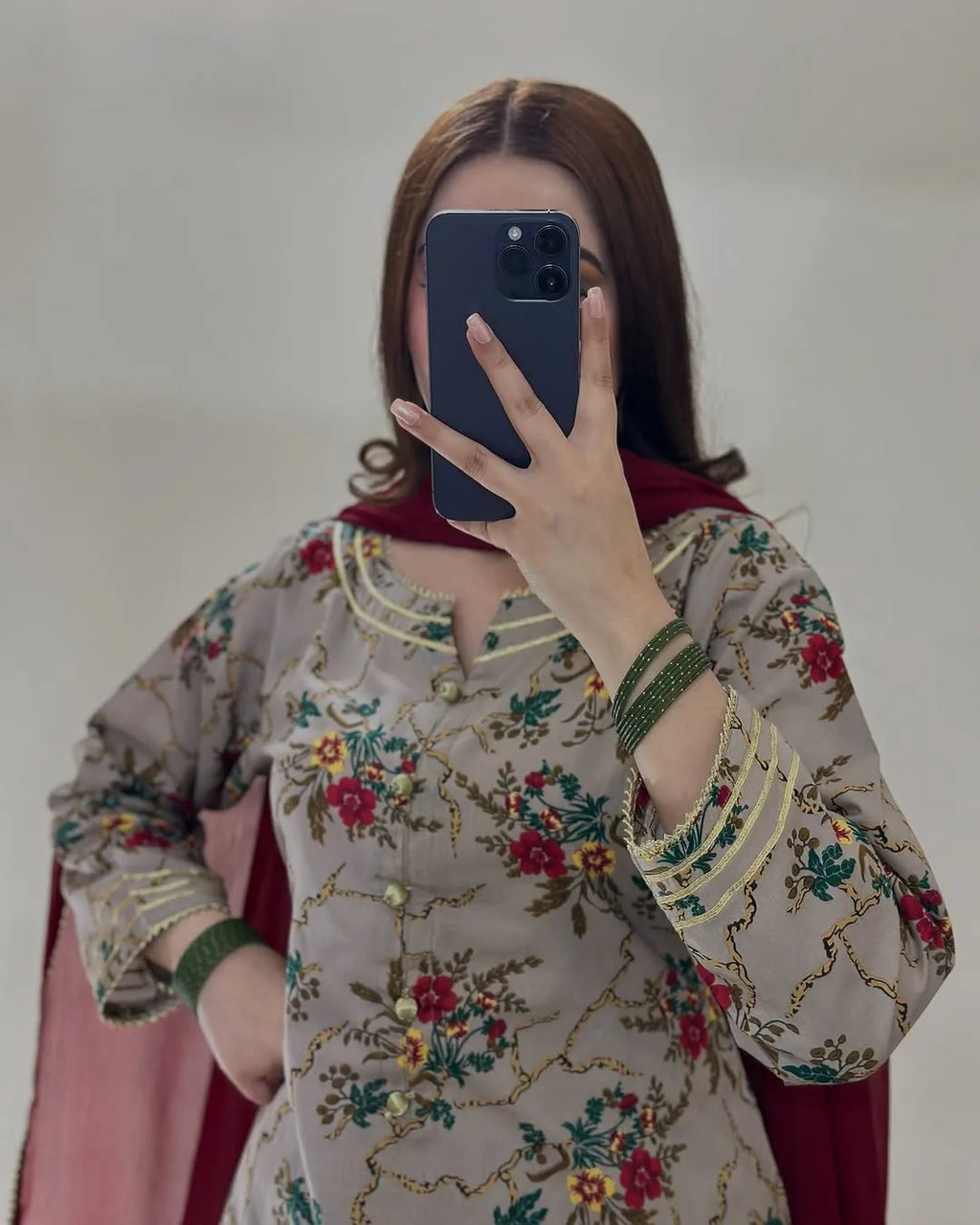 A woman takes a mirror selfie wearing a beige embroidered salwar kameez top with red and green floral patterns, green bangles, and a dark red dupatta.