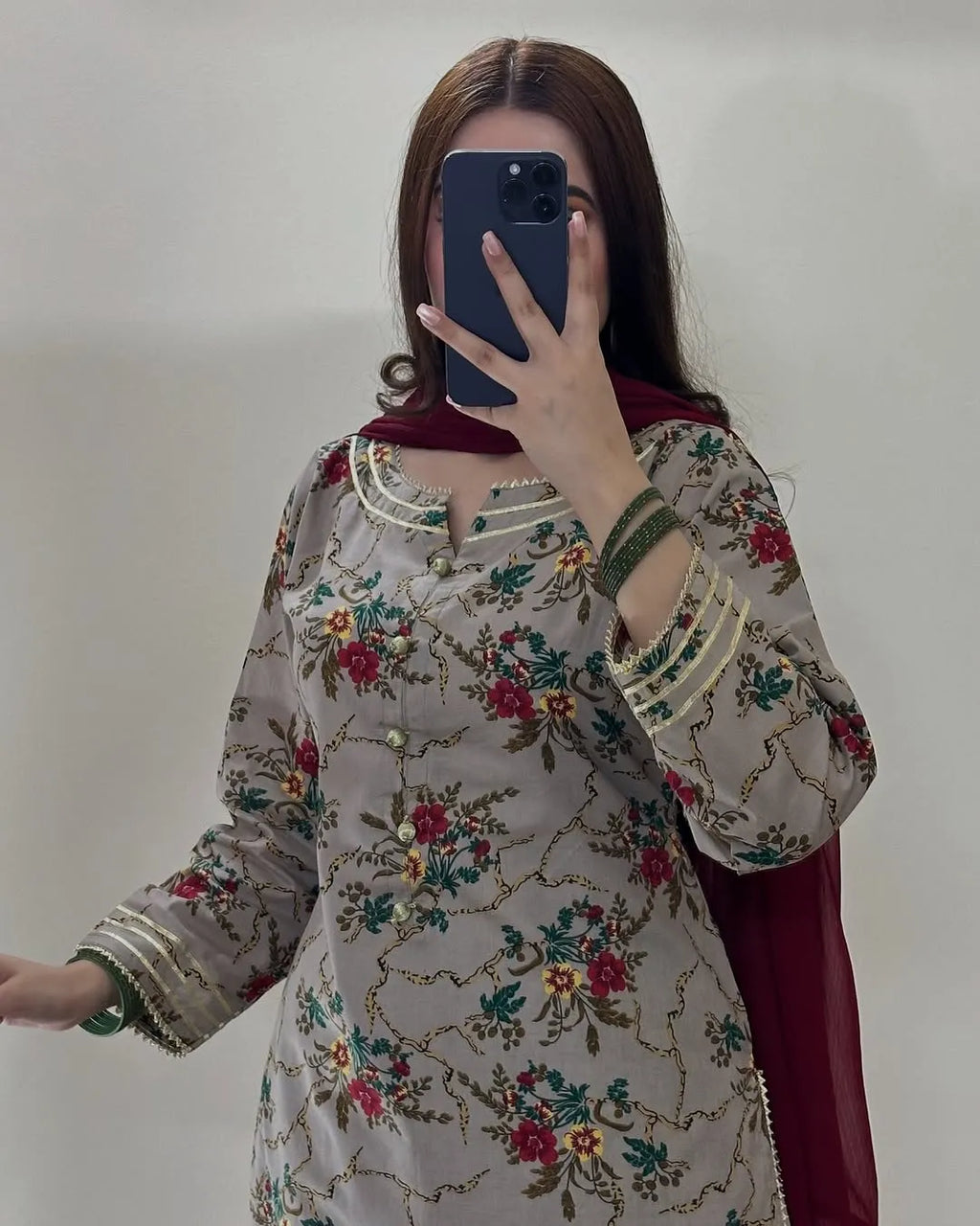 A woman takes a mirror selfie wearing a light beige floral printed kurta with green and red embroidery, a dark red dupatta, and green bangles.