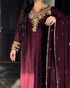 A woman wearing a deep maroon traditional outfit with ornate gold embroidery, a matching embroidered dupatta, and gold jewelry, standing indoors against a dark background.