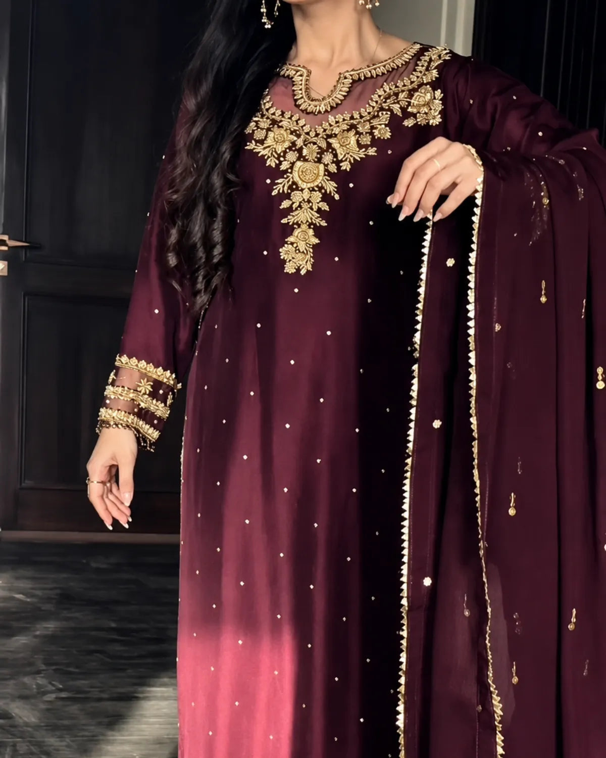 A woman wearing a deep maroon traditional outfit with ornate gold embroidery, a matching embroidered dupatta, and gold jewelry, standing indoors against a dark background.