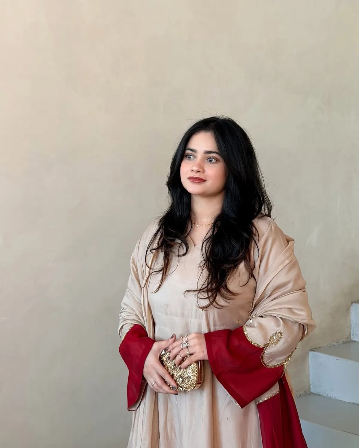A woman with long, wavy black hair is wearing a beige traditional outfit with a deep red shawl draped over her shoulders and holding a small gold clutch. She is standing against a neutral beige plaster wall.