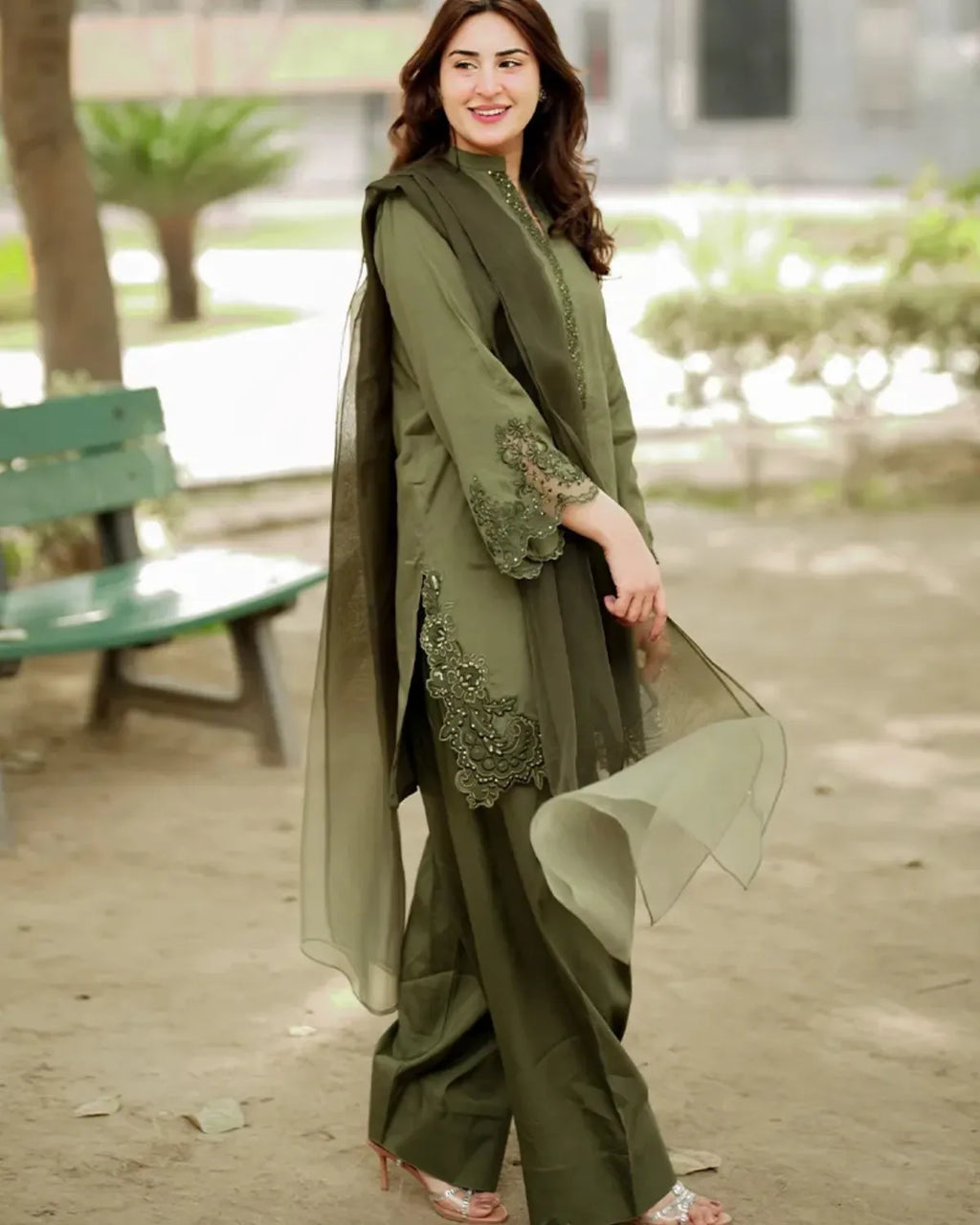A woman wearing an olive green three-piece traditional outfit with matching sheer dupatta and embroidered sleeve details, standing outdoors in a park-like setting.