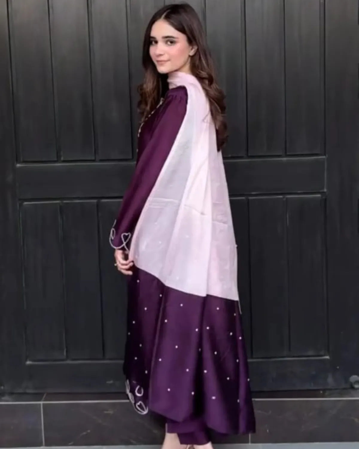 A woman wearing a traditional South Asian outfit with a deep purple skirt decorated with small silver polka dots, a matching purple dupatta, and a light lavender shawl draped over her shoulders, standing in front of a dark paneled door.