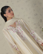 A woman wearing a cream-colored satin traditional outfit with pink and green floral embroidery, paired with a matching dupatta, set against a patterned beige wall.