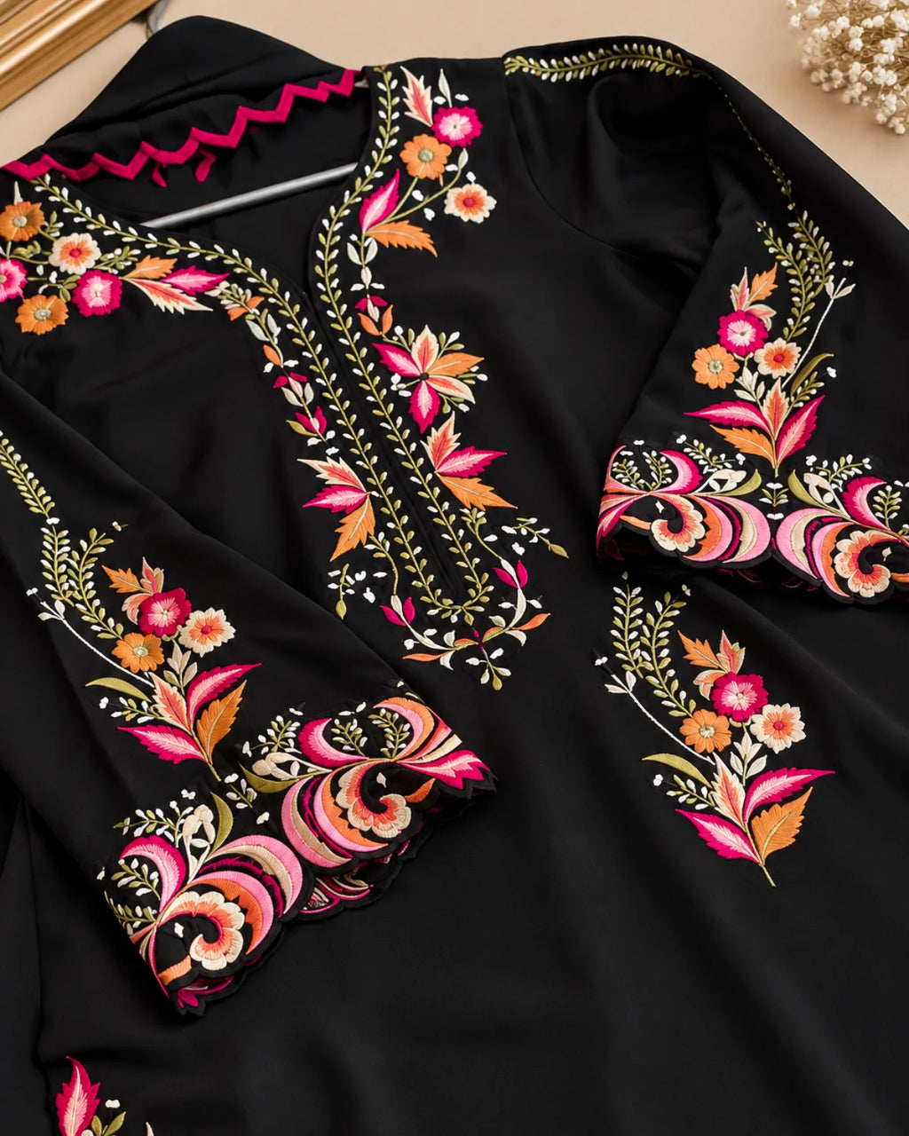 A black long-sleeve tunic or kaftan with elaborate floral and paisley embroidery in pink, orange, gold, and white, shown in a close-up flat-lay view.
