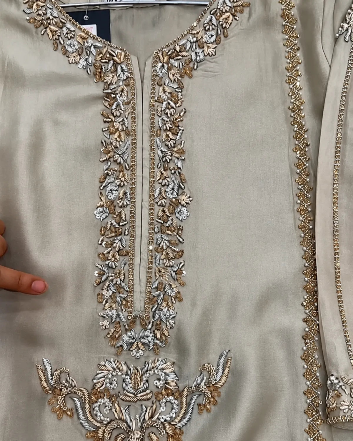Close-up of a beige or champagne-colored embroidered garment, likely a kurta or tunic, featuring ornate metallic silver and gold floral embroidery around the neckline and lower front panel.