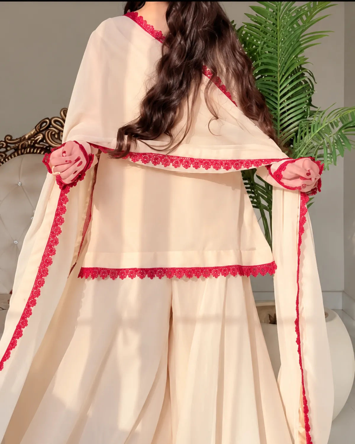 A woman wearing a cream-colored traditional outfit with bright pink-red scalloped borders and matching fabric draped over her shoulders, shown from the back in an indoor setting with a green plant in the background.