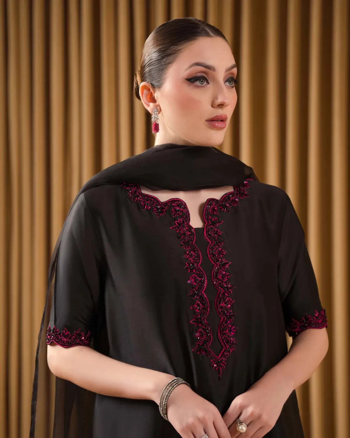 A woman wearing a black short-sleeved tunic with deep V-neckline and sleeve cuff embroidery in dark red, paired with a matching black dupatta, set against a background of gold pleated curtains.