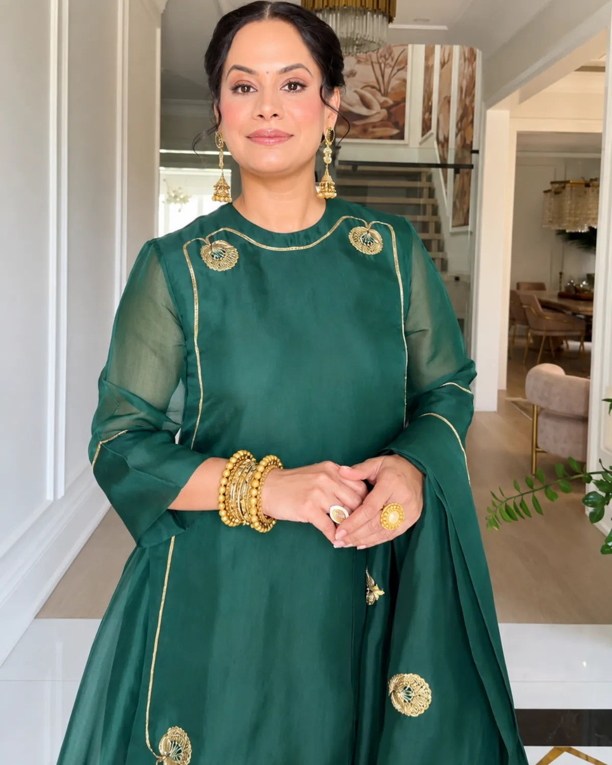 A woman wearing a dark green traditional outfit with gold embroidery, standing in a bright indoor hallway. She is accessorized with gold jewelry, including earrings, bracelets, and rings.