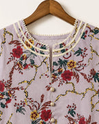 A close-up of a lavender floral printed top hanging on a wooden hanger. The top features a cream-colored collar embellished with small pearls, a matching pearl button placket, and a vibrant floral pattern with red, yellow, and green flowers on a gold vine background.