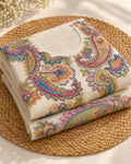 A folded cream-colored cotton kurta featuring vibrant, multicolored paisley and floral embroidery, displayed on a round woven straw placemat with small white flowers in the background.