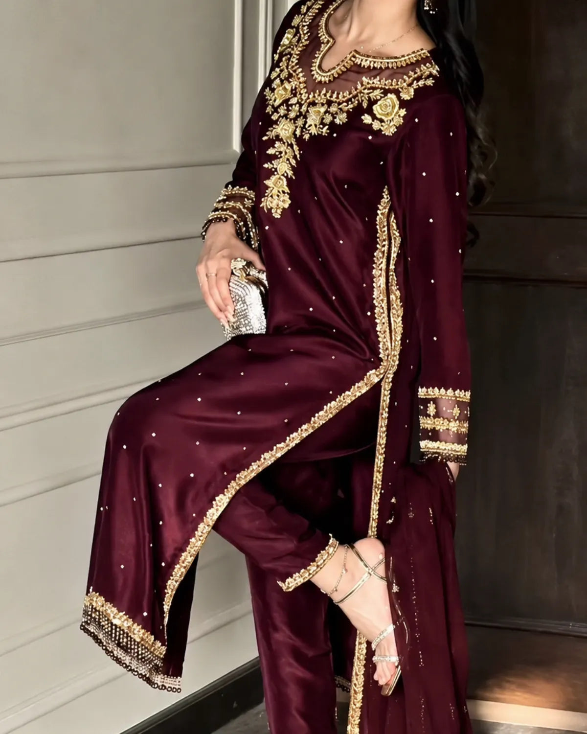 A woman models a deep maroon three-piece ethnic suit with intricate gold embroidery and silver mirror work, paired with a matching dupatta.