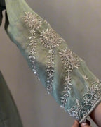 A close-up of a sage green fabric sleeve or garment piece decorated with ornate silver embroidery and beadwork, held by a hand against a plain neutral background.