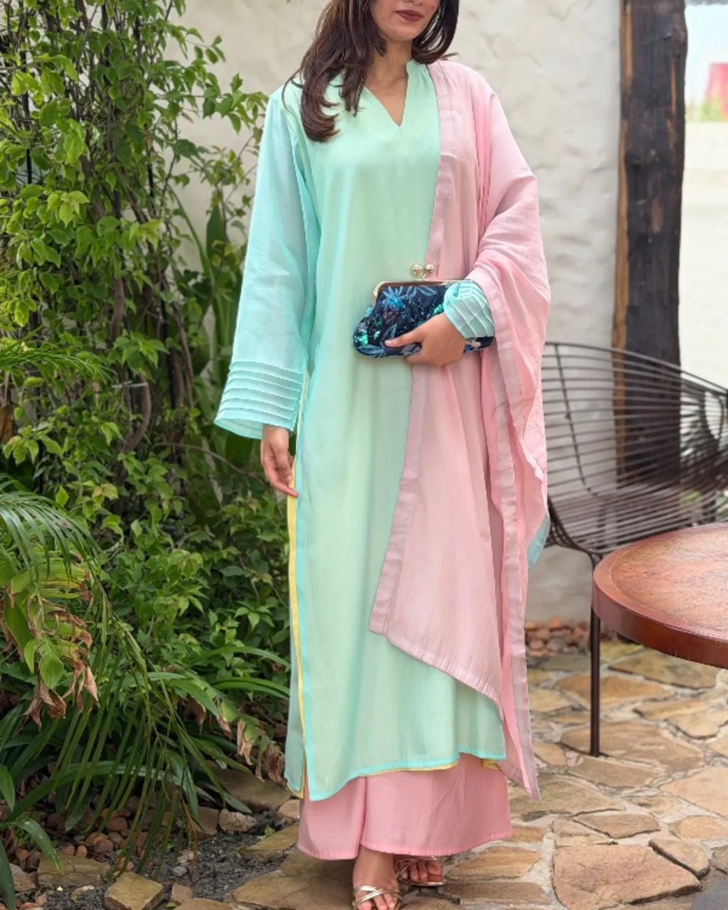 A woman wearing a light mint green and pastel pink traditional outfit with a matching pink dupatta, standing outdoors on a stone patio next to green plants.