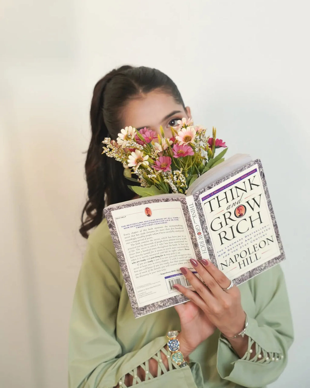 A woman in a sage green top holds an open copy of the book 'Think and Grow Rich' by Napoleon Hill in front of her face, partially obscuring it with a bouquet of pink and white flowers. The background is a clean, off-white wall.