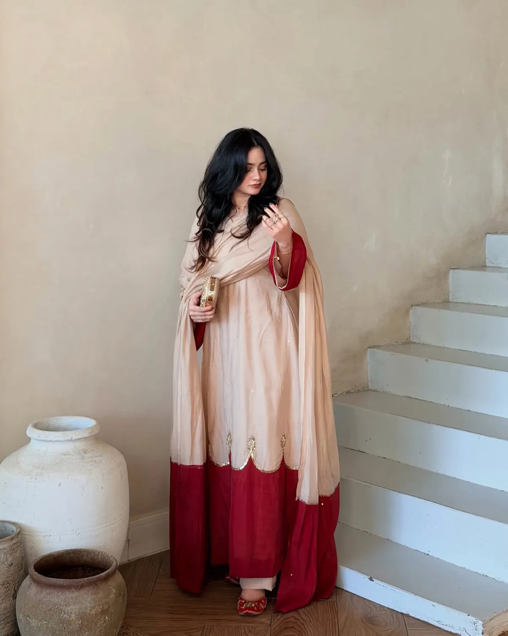 A woman wearing a traditional Indian outfit in beige and deep red stands in a room with beige walls and white stairs. She is holding a small gold object in her hand.