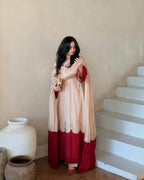 A woman wearing a traditional Indian outfit in beige and deep red stands in a room with beige walls and white stairs. She is holding a small gold object in her hand.