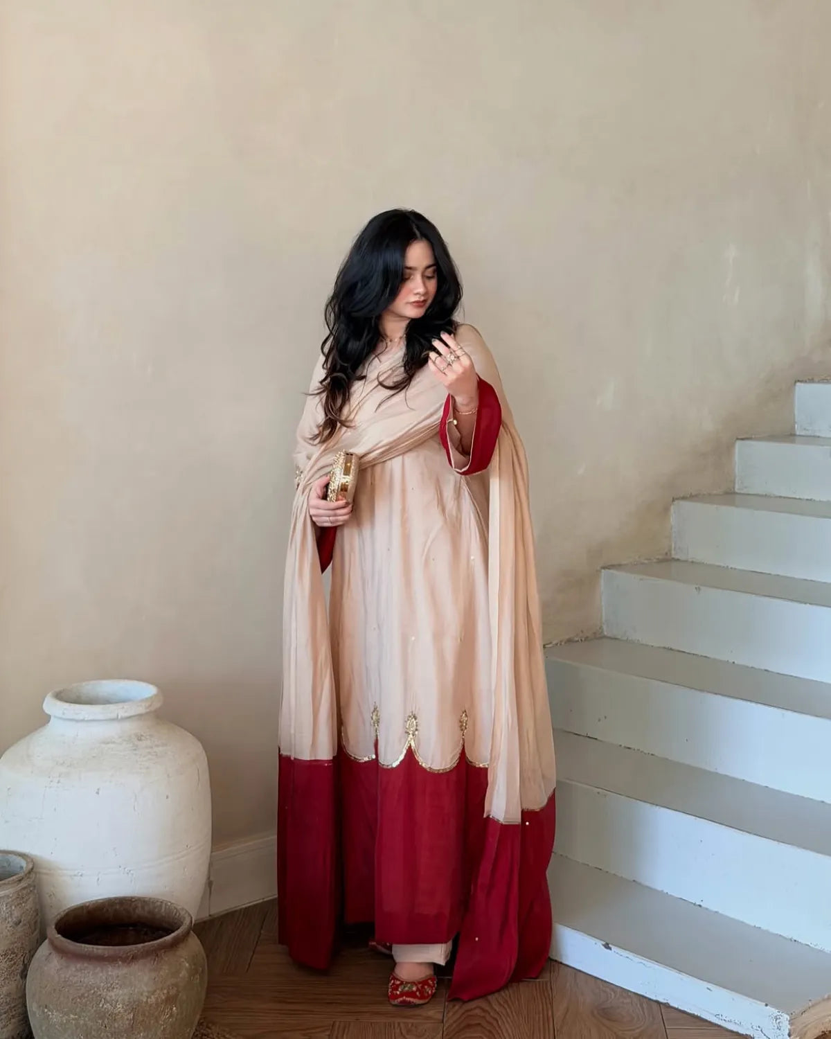 A woman wearing a traditional Indian outfit in beige and deep red stands in a room with beige walls and white stairs. She is holding a small gold object in her hand.