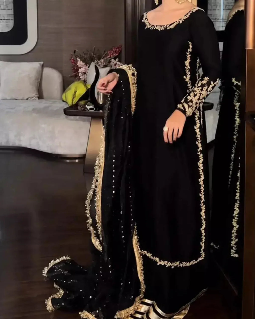 A woman wearing a black traditional outfit with ornate gold embroidery and a matching black dupatta draped over one arm, photographed indoors in a furnished room.