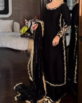 A woman wearing a black traditional outfit with ornate gold embroidery and a matching black dupatta draped over one arm, photographed indoors in a furnished room.