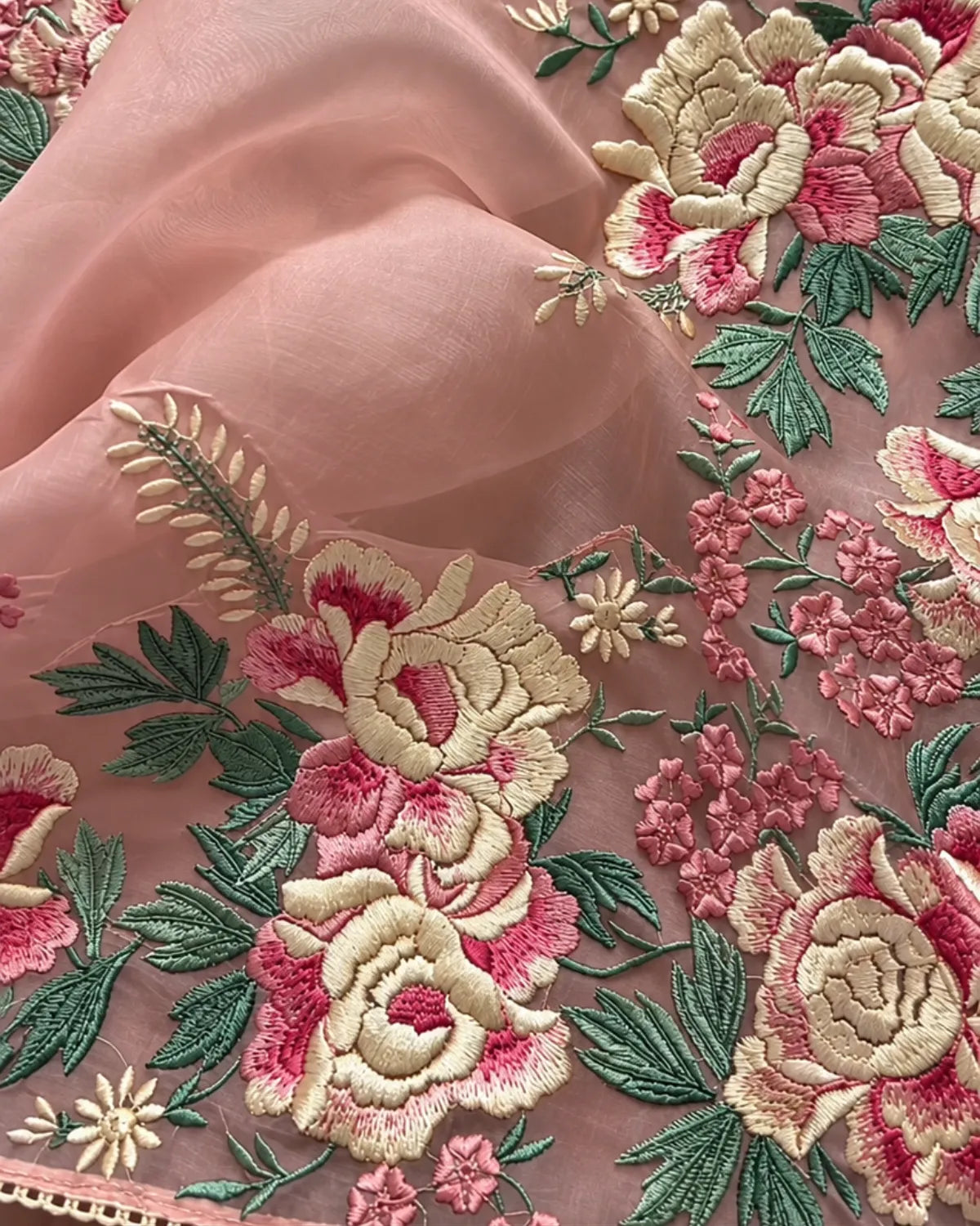 A close-up of peach-pink embroidered fabric featuring large floral motifs in cream, pink, and green, with a sheer matching fabric draped behind it.