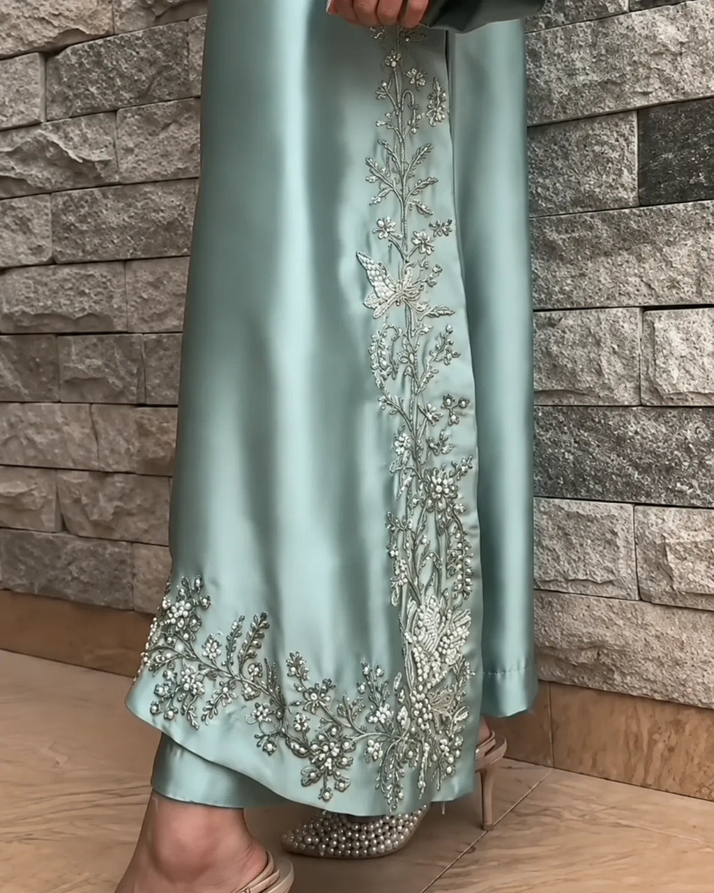 A person wearing a flowing sage green satin abaya with intricate silver and white floral embroidery, paired with silver embellished heels, standing against a gray stone wall.