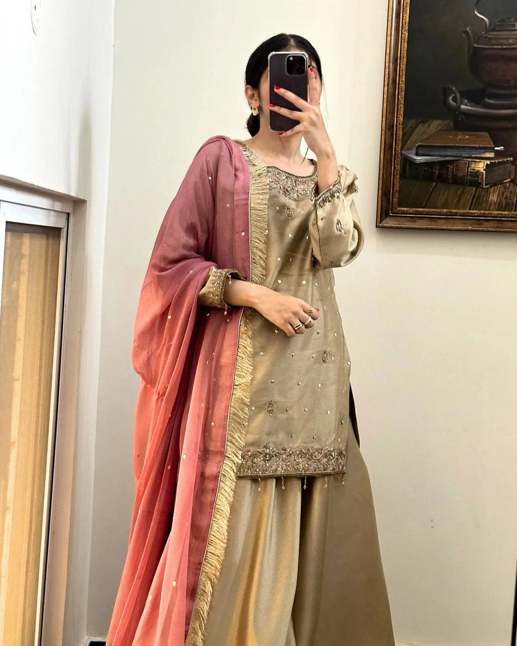 A woman takes a mirror selfie wearing a traditional Indian salwar kameez outfit. The set features a beige embroidered kameez top with gold zari detailing, matching beige wide-leg pants, and a dusty rose gradient dupatta with gold fringe.