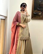 A woman takes a mirror selfie wearing a traditional Indian salwar kameez outfit. The set features a beige embroidered kameez top with gold zari detailing, matching beige wide-leg pants, and a dusty rose gradient dupatta with gold fringe.