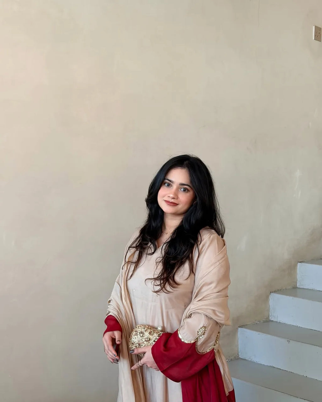 A woman with long dark hair stands indoors against a beige wall and white stairs, wearing a light beige traditional outfit with deep red sleeves and holding a small gold patterned clutch.