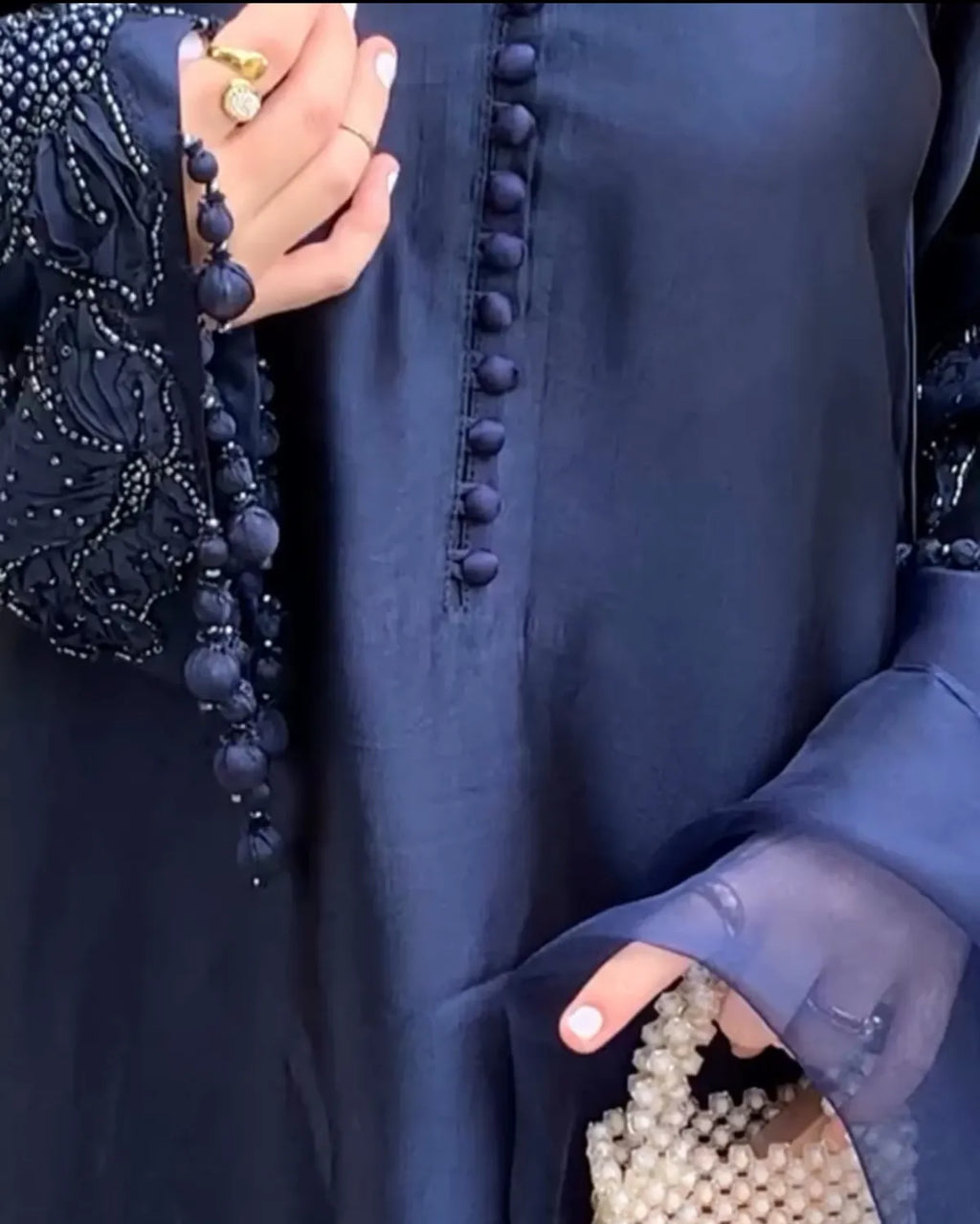 Close-up of a navy blue abaya dress with decorative beaded trim and a central row of pom-pom details, shown on a model holding a pearl beaded clutch.