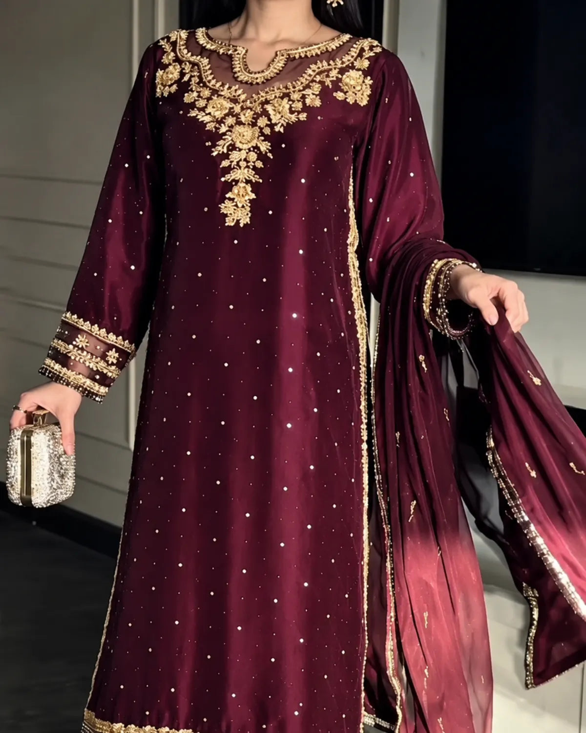 A woman wearing a deep maroon traditional outfit with extensive gold embroidery on the neckline, sleeves, and hem, paired with a matching sheer dupatta and holding a small metallic clutch.