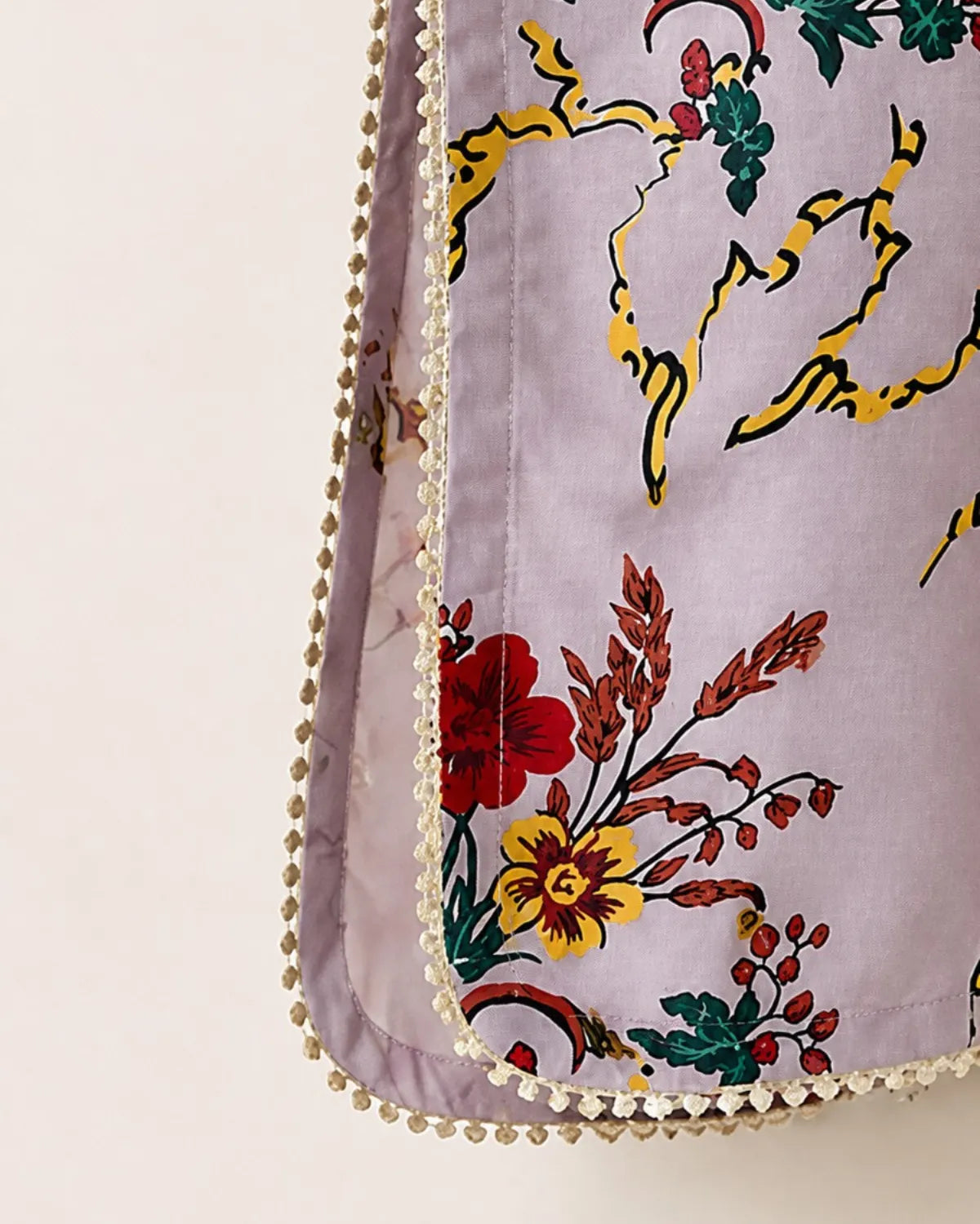 Close-up of a lavender fabric item with a colorful floral print in red, yellow, and green, finished with a decorative cream pom-pom trim.