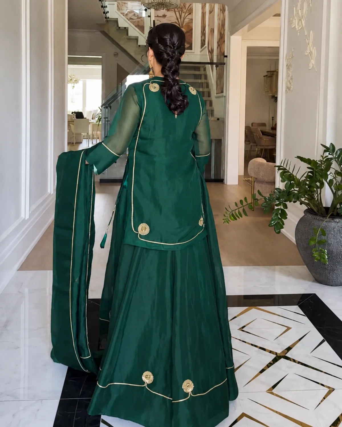 A woman viewed from behind wearing a dark green traditional outfit with gold trim and a matching dupatta, standing in a modern indoor hallway with a staircase and potted plants in the background.