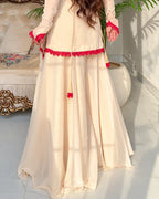 A woman wearing a cream-colored traditional outfit with bright red lace trim and tassel details, standing in a sunlit indoor setting with a sofa and potted plants in the background.