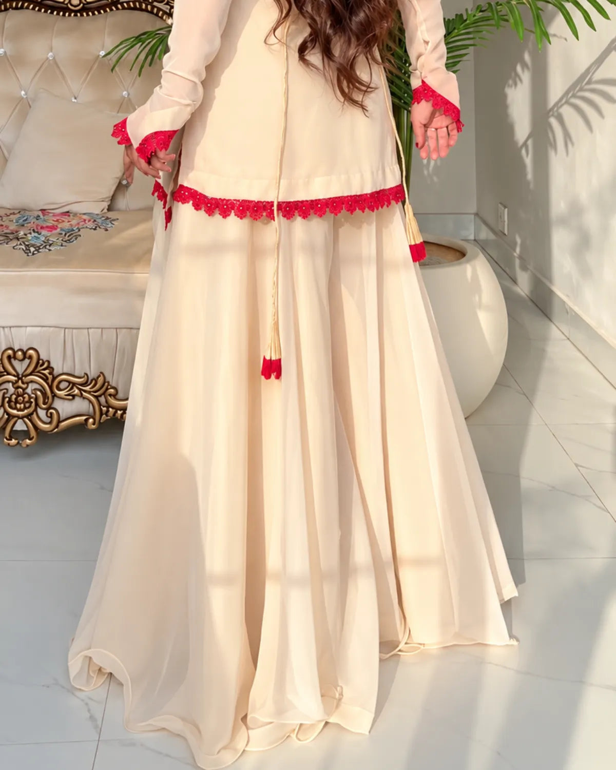 A woman wearing a cream-colored traditional outfit with bright red lace trim and tassel details, standing in a sunlit indoor setting with a sofa and potted plants in the background.