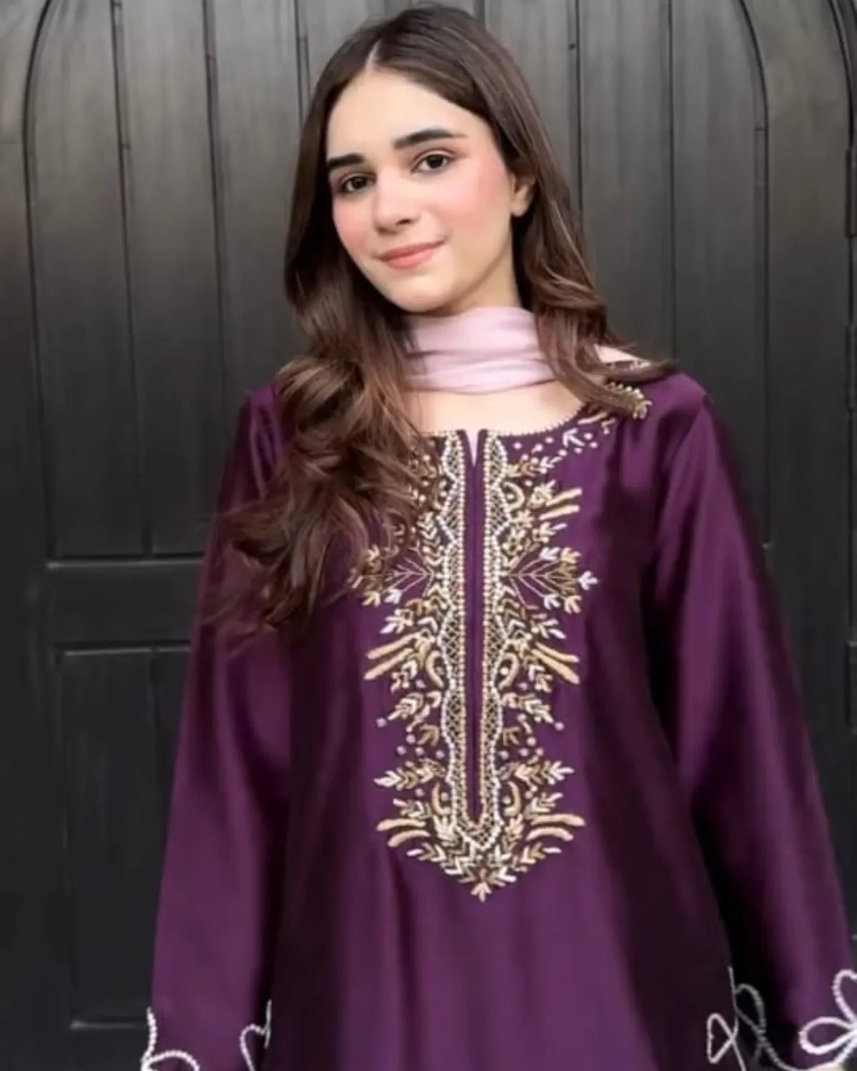 A young girl with long brown hair wearing a purple long-sleeve tunic with gold and white embroidery on the chest, paired with a light pink neck scarf, standing in front of a dark wooden door.