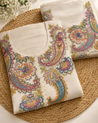 Two folded off-white embroidered kurtis with colorful paisley and floral motifs, arranged on a round woven jute mat with small white flowers in the corner.