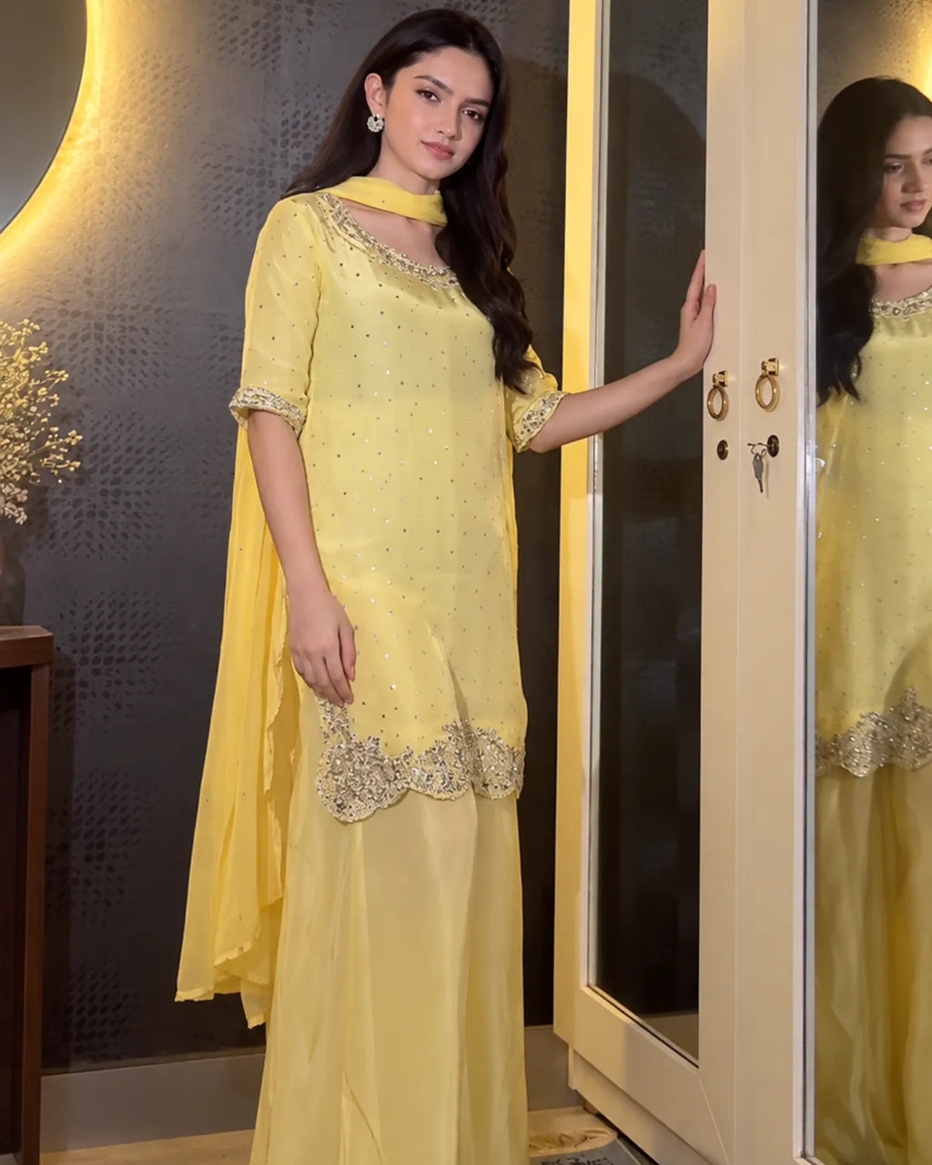 A woman models a light yellow traditional outfit with silver embellishments and a matching dupatta in an indoor setting with a mirror reflecting her profile.