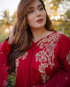 A woman wearing a deep red traditional embroidered outfit with intricate gold and beige floral embroidery on the neckline and sleeves, photographed outdoors with a soft blurred background.
