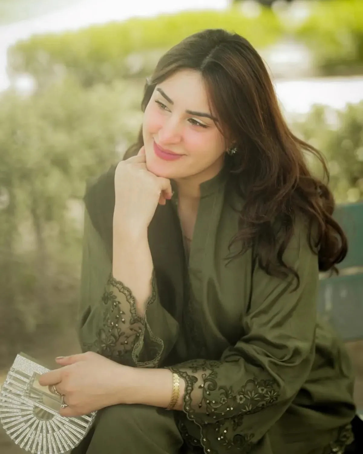 A woman with long dark brown hair is shown in a lifestyle setting, wearing an olive green traditional outfit with black lace embroidery on the sleeves. She is holding a silver folding fan and resting her chin on her hand, with a soft-focus green outdoor background.