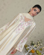 A woman wearing a cream satin abaya with pink and green floral embroidery, posed against a patterned beige wall with decorative props at the bottom right.