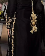 A close-up of a black traditional dress with ornate gold embroidery along the sleeve and side, featuring a large dangling gold embellishment on the chest.
