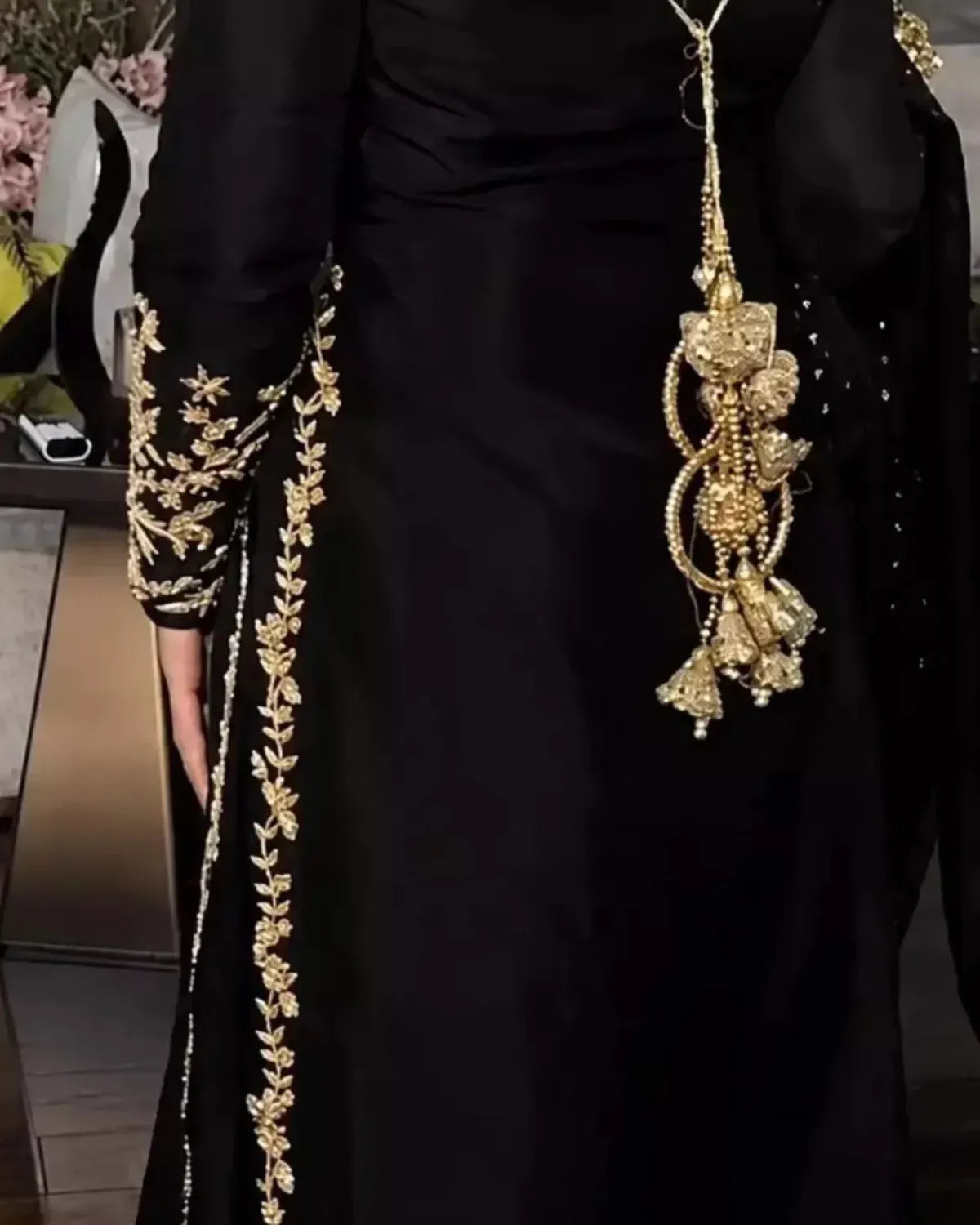 A close-up of a black traditional dress with ornate gold embroidery along the sleeve and side, featuring a large dangling gold embellishment on the chest.