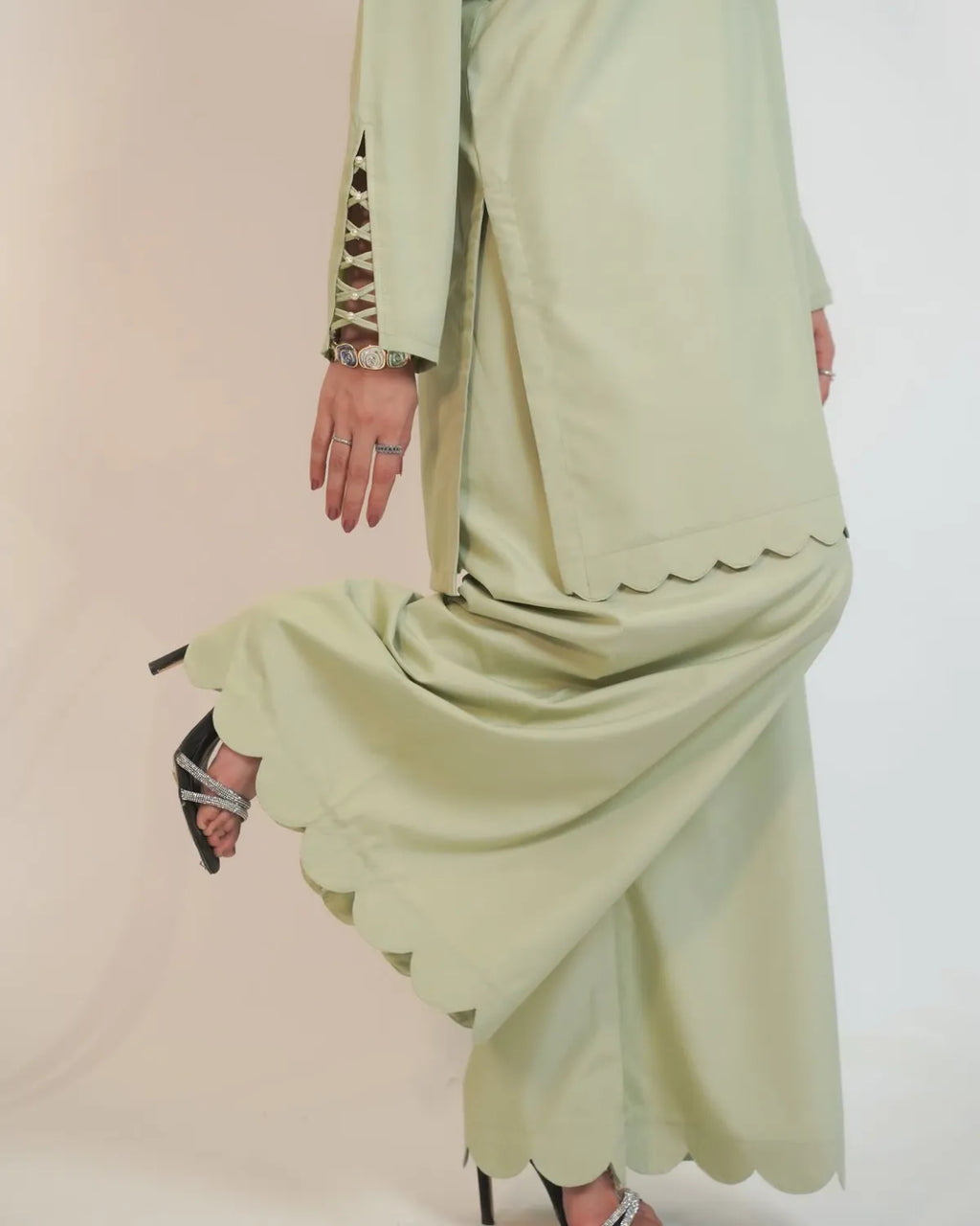 A model wearing a light sage green traditional outfit with scalloped edges and lace-up sleeve details, paired with black high-heeled sandals against a plain light background.