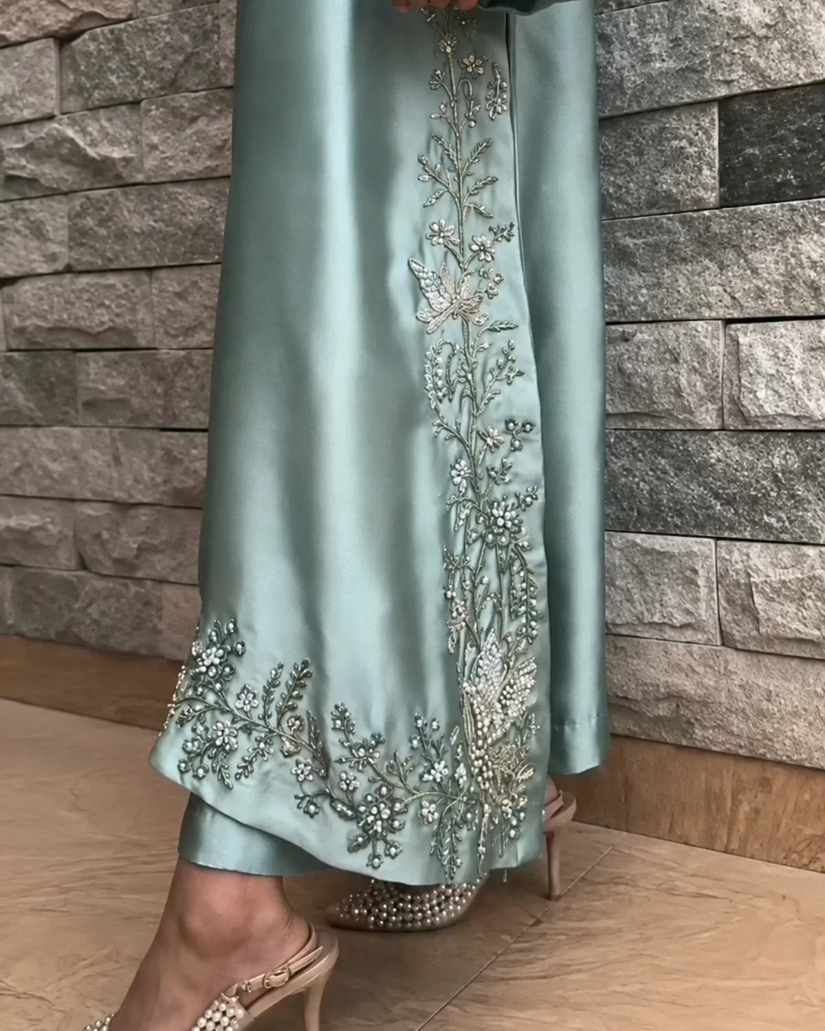 A close-up of a person wearing a flowing sage green satin dress with intricate silver and white floral embroidery along the side and hem, paired with embellished heels, standing in front of a gray stone wall.